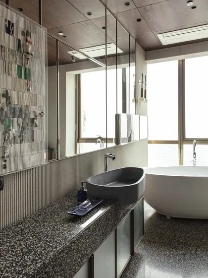 A view of the daughter's bathroom, highlighting the Gomaads concrete wash basin set against a terrazzo counter. Mirrored cabinets expand the space and reflect the natural light from the large window.