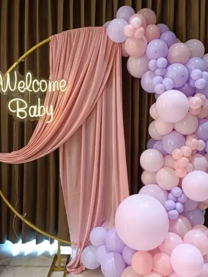 A soft and dreamy welcome for a new baby. I used a circular gold frame draped with delicate pink fabric, a 'Welcome Baby' neon sign, and a beautiful balloon arch in pastel pinks and purples to create this gentle and inviting atmosphere.