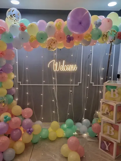 A soft and cheerful welcome home setup using a pastel rainbow of balloons. The arch is complemented by a "Welcome" neon sign, fairy lights, and 'BABY' blocks.