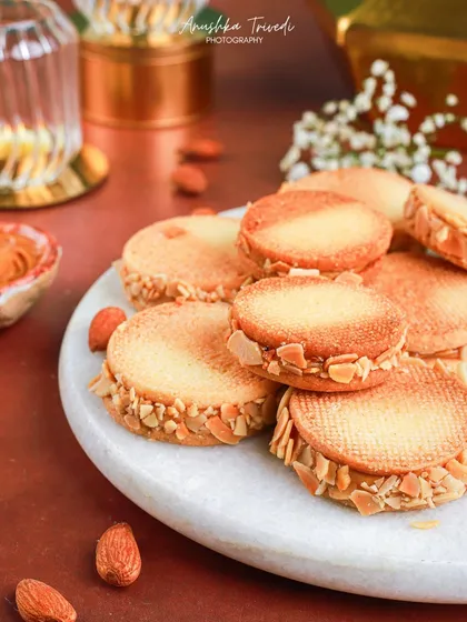 A plate of almond sandwich cookies, styled with a warm, moody background. The lighting creates a soft glow that feels perfect for an evening Diwali celebration.