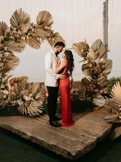 A breathtaking portrait of the couple standing on a rustic stone platform, framed by an arch of dried pampas grass and florals. The bride's red gown provides a stunning pop of color against the neutral decor.
