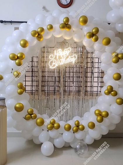 A classic and elegant circular balloon ring in white and gold, with a silver fringe backdrop and a "Happy Birthday" neon sign.