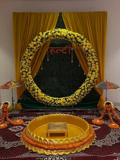 A beautiful haldi setup featuring a large circular arch of yellow flowers against a green backdrop. The lotus seating area is placed on a traditional carpet.