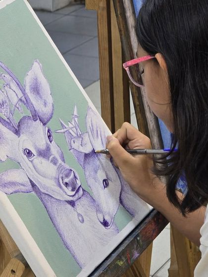 A close-up of a young artist's hand as she adds fine details to the antlers of a deer in her ballpoint pen illustration.