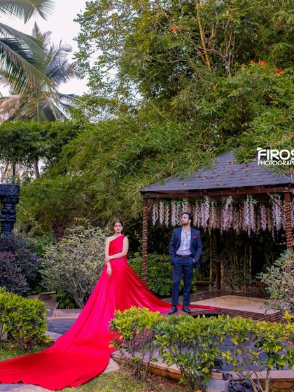 A wide shot of a couple on the wooden bridge, with the flowing red dress creating a stunning visual against the green garden.
