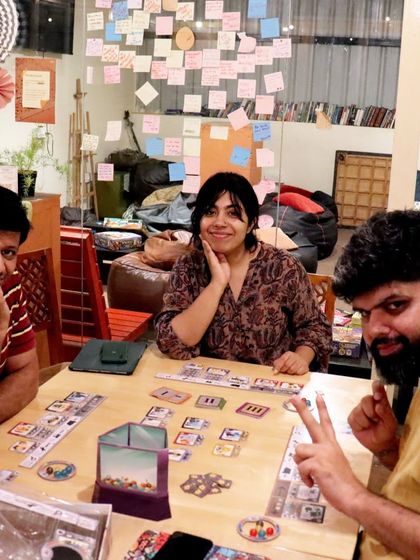 Three players are focused on a game of 'Vinhos' at a table near our wall of love, covered in sticky notes from our community.