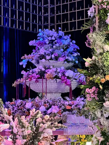 A towering floral masterpiece, blending lush blooms in vibrant hues. This installation, illuminated with dramatic lighting, serves as a show-stopping backdrop for any celebration. 💜
