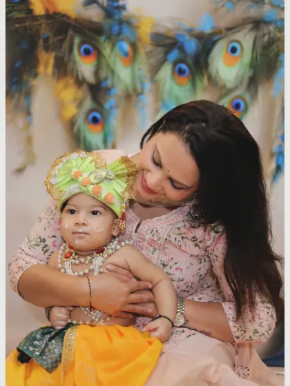A loving portrait of a mother holding her baby, who is dressed in a Krishna costume against a backdrop of peacock feathers.