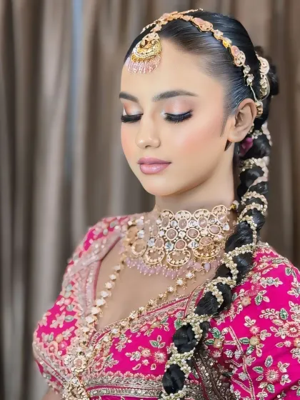 A close-up of a bride in a vibrant pink lehenga. The makeup is soft and romantic, with pink tones on the eyes and cheeks, and a sleek, traditional braided hairstyle.