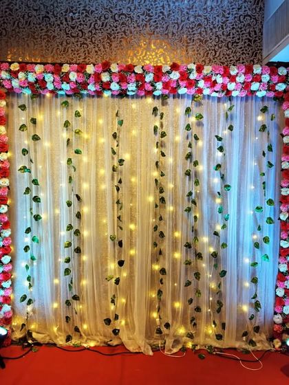 A simple yet beautiful backdrop for a home Mehndi function. We create a frame of red and pink roses around a sheer curtain decorated with fairy lights and hanging green vines.