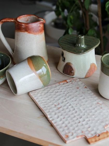 A charming set of student pieces including a pitcher, a house shaped lidded pot, and a textured platter, all in earthy green and white glazes.