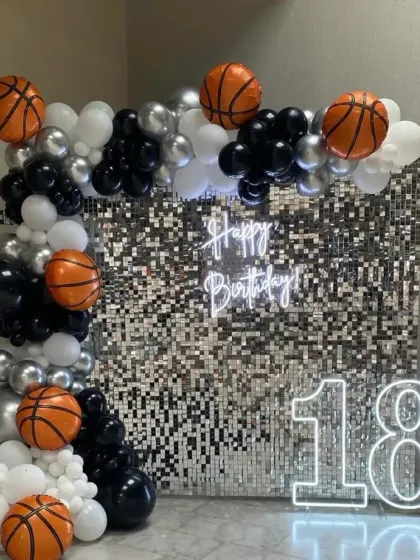 A modern and stylish 18th birthday setup. This features a shimmering silver sequin wall, a custom "Happy Birthday" neon sign, and a large "18" marquee number, all framed by a balloon arch in black, white, silver, and basketball-themed balloons.