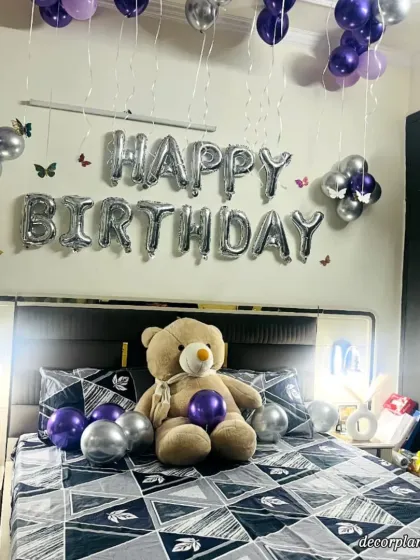 A beautiful bedroom surprise featuring a teddy bear on the bed, surrounded by purple and silver balloons. The 'Happy Birthday' foil letters and butterfly decals on the wall complete this sweet and thoughtful gesture.