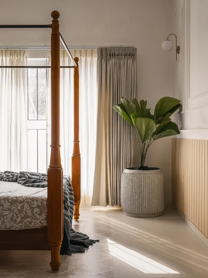 Natural light filters through sheer curtains, illuminating a corner of the bedroom. The space is kept simple with a fiddle-leaf fig plant and minimalist wall paneling to complement the traditional bed frame.
