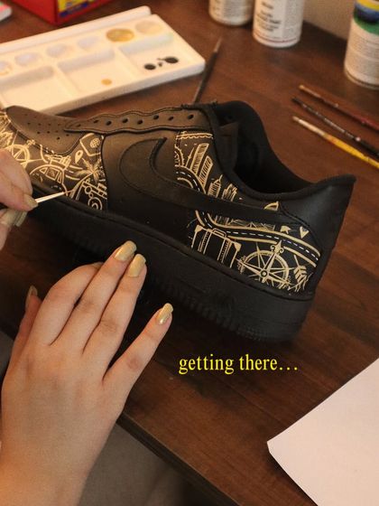 One brushstroke at a time. Here's a look at the process of painting a detailed city skyline and travel-themed icons onto a pair of black Nike Air Force 1s.