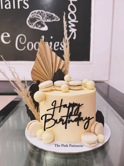 A modern cake in neutral tones, decorated with macarons, dried pampas grass, and a black "Happy Birthday" script.