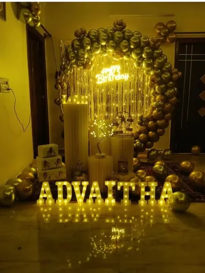A glamorous gold and black birthday decoration for Advaitha. The room is lit by a neon sign and large marquee letters, with a balloon ring and fairy lights creating a warm, celebratory glow.