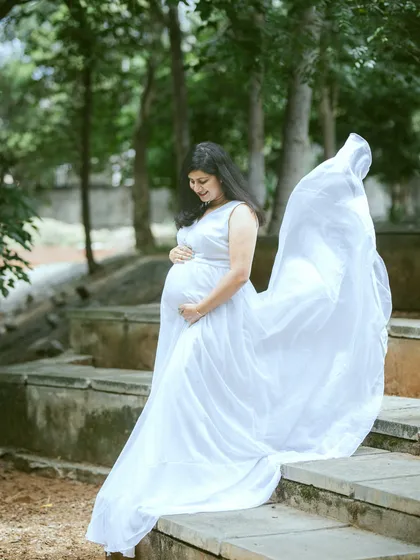 An expectant mother looking serene in a flowing white gown during an outdoor maternity session. The natural park setting and the gentle toss of the fabric create a timeless and angelic portrait.