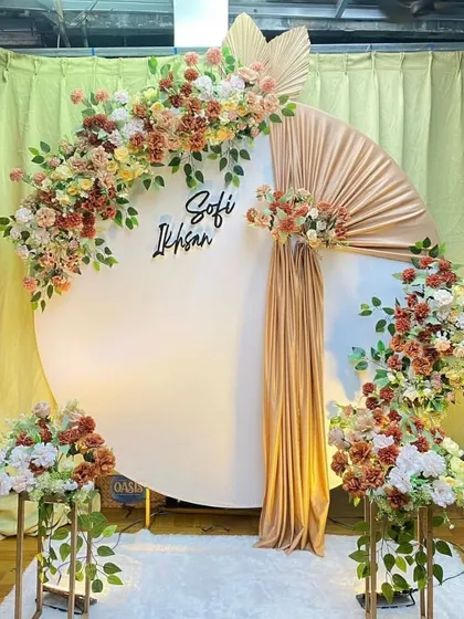 A stylish engagement backdrop with a large white circular panel and earthy-toned floral arrangements. A touch of gold fabric adds a bit of shimmer, and the couple's names can be added for a personalized touch.