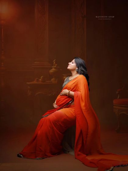 A painterly portrait of a mother-to-be seated in a gracefully draped orange saree. The side lighting creates a dramatic and artistic effect.
