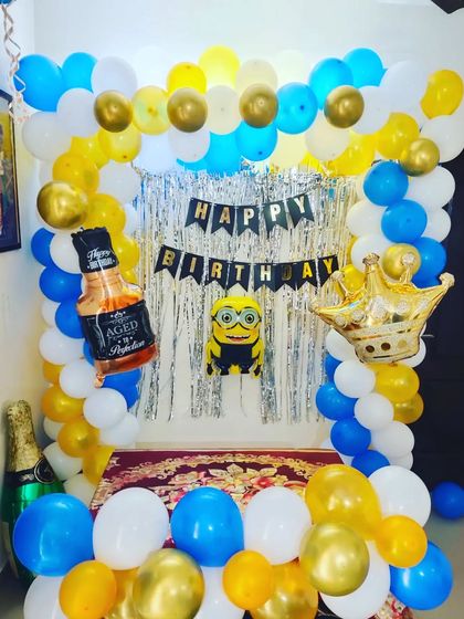 A fun Minions-themed birthday party decoration. The setup includes a blue, yellow, and white balloon arch against a silver tinsel curtain, with Minion, crown, and whiskey bottle foil balloons for a playful touch.