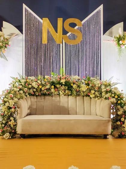 A centered, close-up view of the couple's seating, nestled within an arch of pastel-colored roses and hydrangeas. The custom 'NS' monogram and shimmering backdrop create a personalized and picture-perfect setting.