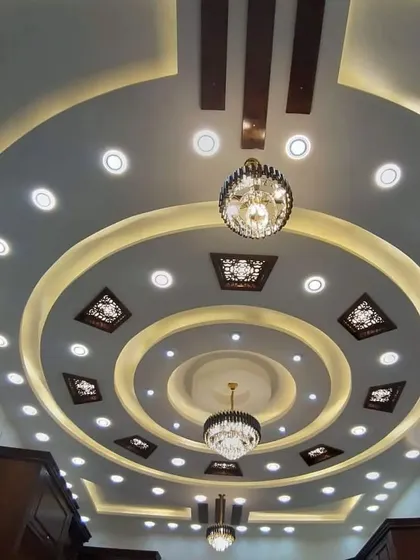 A grand, multi-ring ceiling designed for a large hall. The numerous spotlights and two chandeliers ensure the space is brilliantly lit, highlighting the intricate POP work.