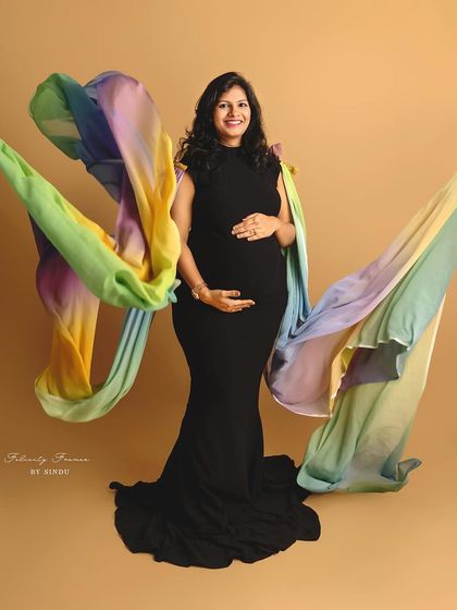 A smiling mother-to-be in a classic black gown, with a vibrant, rainbow-colored fabric flowing around her like wings. This creative studio shot celebrates the miracle of life.