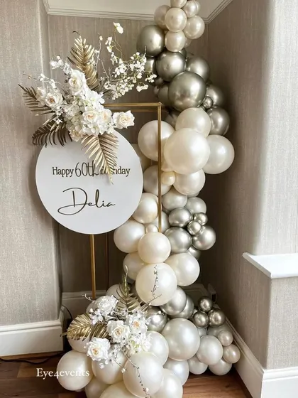 This elegant corner decoration was created for a 60th birthday. It features a custom "Happy 60th Birthday" sign on a gold stand, framed by a beautiful balloon garland in white, silver, and cream, with delicate floral accents.