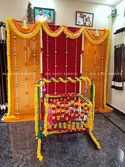 A classic and traditional cradle ceremony decoration for a home event. The setup uses a rich combination of red and orange drapes, with garlands of marigold and white flowers. The baby's cradle is the main focus, heavily decorated with matching yellow and red flowers to create a warm and auspicious atmosphere.