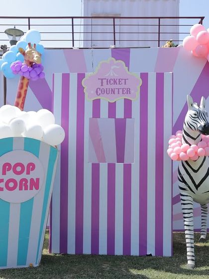 A whimsical ticket counter and popcorn stand for an outdoor carnival. This setup features a giant popcorn box prop, a life-sized zebra wearing a balloon necklace, and a striped ticket booth, creating a perfect photo opportunity.