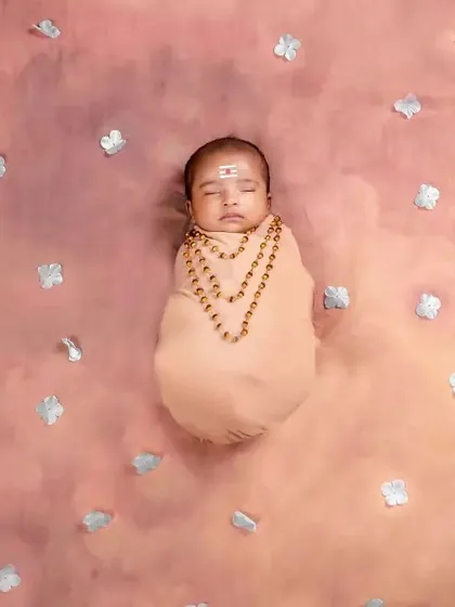 A newborn styled as a little devotee for Vinayagar Chathurthi, swaddled and lying on a soft, cloud-like background with silver petals.