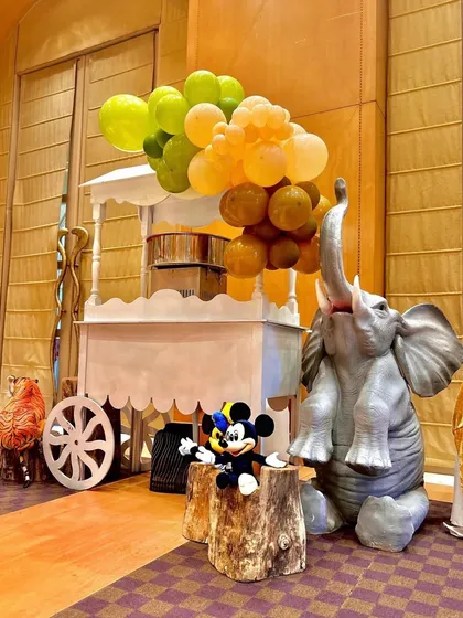 A whimsical corner of the jungle safari party, where a life-sized elephant prop and a Mickey Mouse plush sit next to a cart decorated with an organic balloon garland in shades of green and orange.