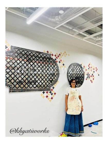 Standing with the completed wall art in a corporate office. The design includes large, mirrored traditional motifs surrounded by a scattering of plus-sign patterns, creating a dynamic and reflective surface.