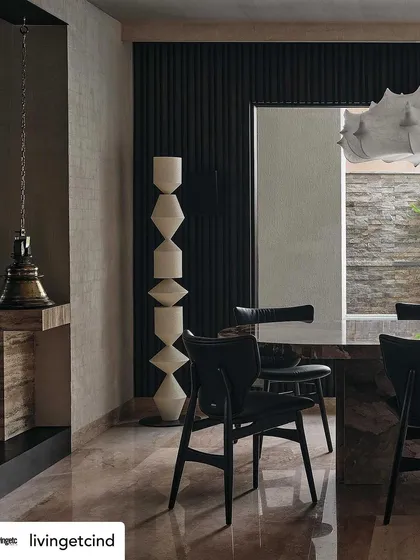 A detail from the dining area, showing the heavy stone table, modern black chairs, and a sculptural white floor lamp. The hanging temple bell adds a touch of Indian heritage.