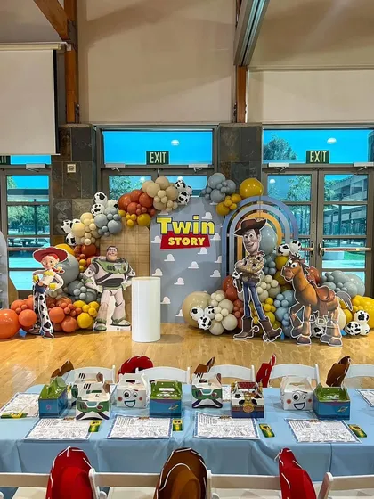 A "Twin Story" birthday party based on the movie Toy Story. The backdrop features Woody, Buzz Lightyear, and Jessie, with cow-print balloons and themed table settings.