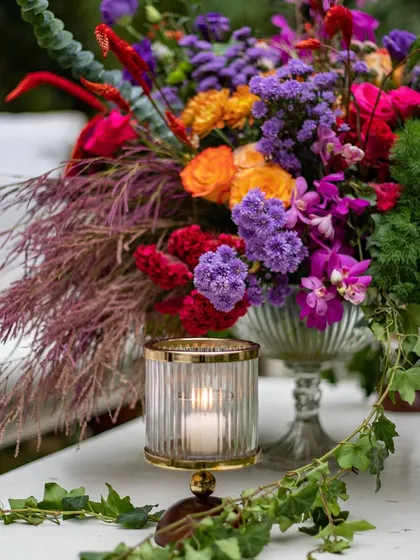 A vibrant floral centerpiece designed for a garden soirée. The arrangement features a bold mix of purples, oranges, and reds, complemented by wild greenery and the soft glow of a candle, perfect for outdoor event styling.