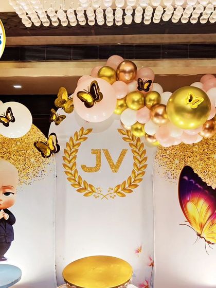 A glamorous joint celebration featuring Boss Baby and Butterfly themes. The decor includes gold and pink balloons, character cutouts, and a custom 'JV' monogram.