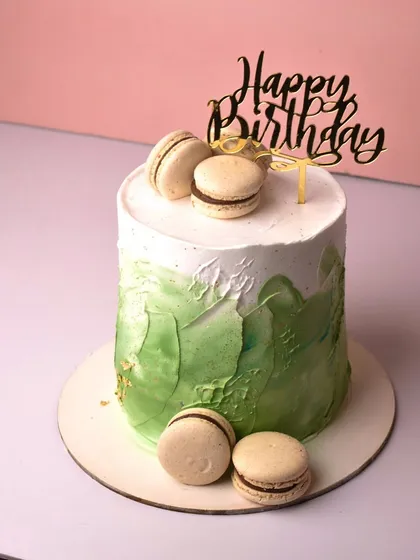 Name a better duo than cake and macarons. This tall cake features a modern green watercolor or spatula-painted effect, topped with a "Happy Birthday" sign and delicious chocolate-filled macarons.