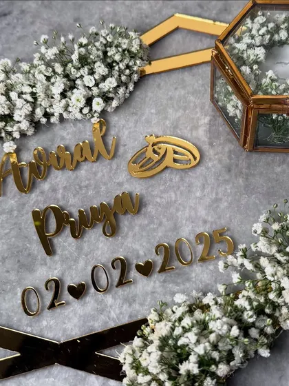 A close-up of the minimalist acrylic frame platter. The focus is on the beautiful gold script of the couple's names and the delicate baby's breath in the ring box.