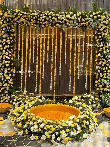A luxurious Haldi setup using real flowers. The square frame is covered in marigolds and white chrysanthemums, with a flower-filled urli at the center for a truly opulent look.