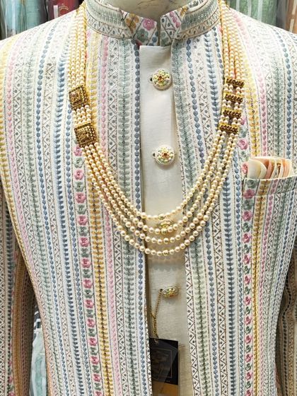 A close-up of a modern Indo-Western sherwani. The jacket features subtle multi-color striped embroidery, layered over a cream kurta and accessorized with a five-layer pearl mala.
