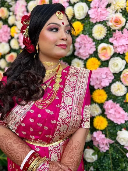 This beautiful bride is ready for her reception. Her makeup features a glamorous pink and brown smokey eye, paired with a traditional bindi and intricate gold jewelry against a lovely floral backdrop.