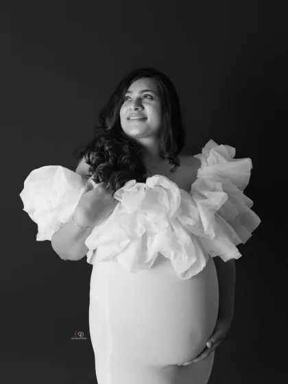 A timeless black and white studio portrait that radiates elegance. The dramatic ruffles of the gown beautifully frame the baby bump, creating a classic and powerful image of motherhood.