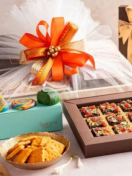 A beautifully assembled Diwali hamper featuring a large wrapped gift box, jars of cookies, a box of date squares, and other festive treats. The vibrant orange ribbon adds a touch of festive cheer to this generous gift.