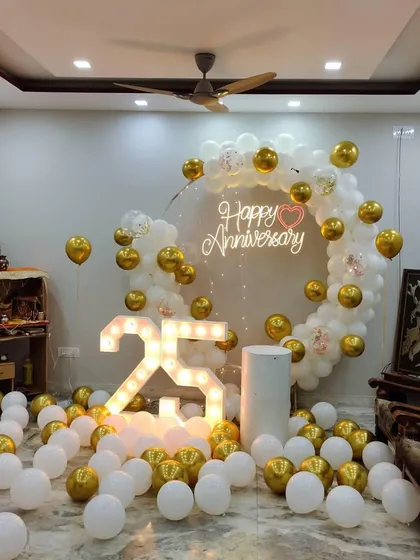 An elegant 25th-anniversary decoration in a living room. A white and gold balloon ring surrounds a neon sign, with a large light-up '25' and balloons scattered on the floor.