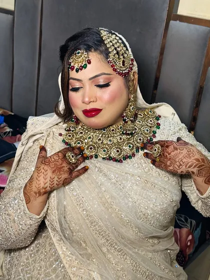 This bride is adjusting her heavy necklace, giving a glimpse of her complete Nikkah look. The makeup, with its radiant base and bold red lip, stands out beautifully against her cream-colored, embellished outfit.