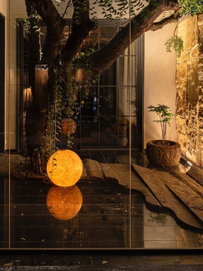 A night view of the KEUS courtyard, where a glowing spherical lamp is reflected on the polished black granite floor. The lighting design creates a magical and contemplative mood, highlighting the texture of the stone wall and the form of the tree.