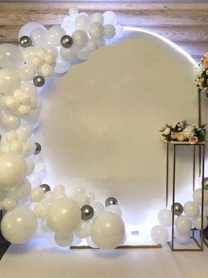 A modern and chic baptism decoration with a backlit circular backdrop. The setup is framed by a white and silver balloon arch and accented with floral bouquets.