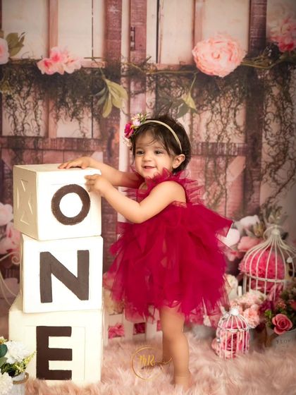 A proud toddler stands next to "ONE" blocks during her first birthday shoot, surrounded by pink flowers.